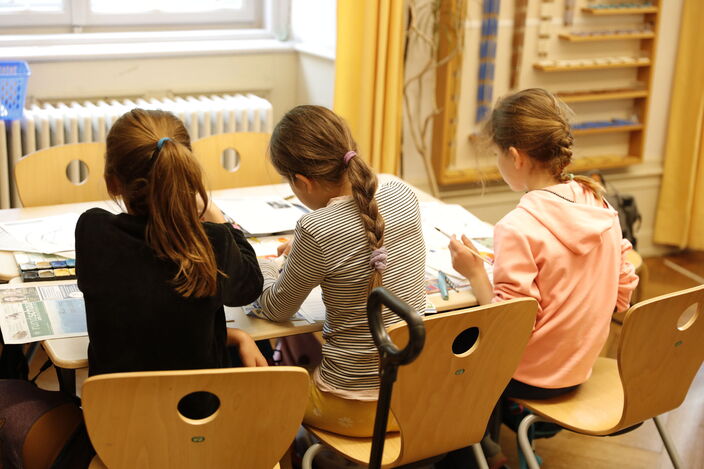 Grundschule Impressionen mit Mathematerial im Hintergrund und drei Schülerinnen zusammen